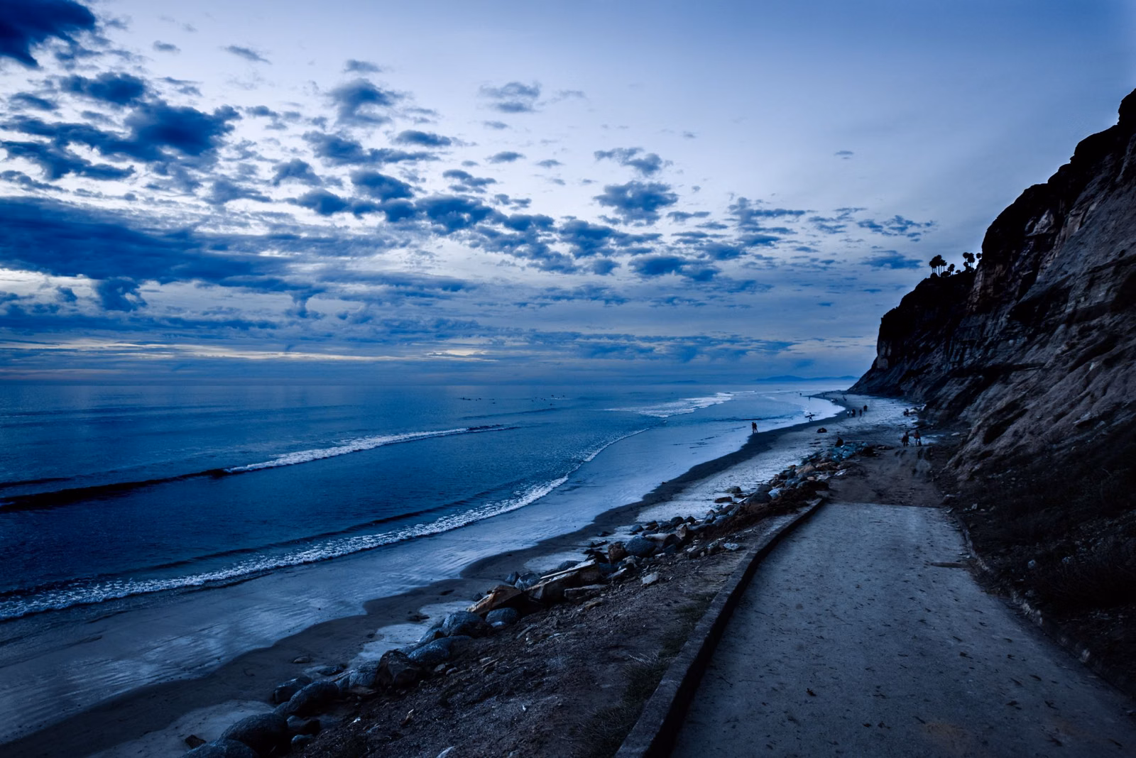A sandy path along the base of coastal cliffs beside the surf under evening clouds